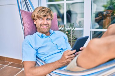 Orta yaşlı yakışıklı bir adam evinin terasında, bir hamakta dokunmatik bir aletle dinleniyor.