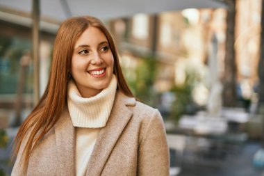 Genç kızıl kız gülümsüyor, mutlu bir şekilde şehirde duruyor..