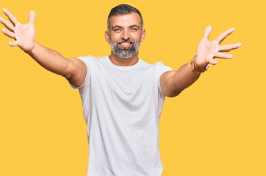 Orta yaşlı, beyaz tişört giyen yakışıklı bir adam kameraya sarılmak için gülümseyerek bakıyor. Mutluluğu kucaklayan neşeli bir ifade.. 