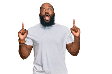 Gündelik beyaz tişört giyen Afro-Amerikalı genç adam şaşırmış ve şaşırmış bir şekilde yukarı baktı ve parmaklarıyla işaret ederek kollarını kaldırdı.. 
