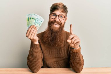 Genç İrlandalı kızıl saçlı adam elinde 5 Singapur Doları banknotlarla masaya oturmuş bir fikir veya soruyla gülümsüyor ve mutlu bir yüzle bir numarayı işaret ediyor. 