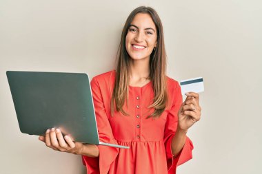 Dizüstü bilgisayar ve kredi kartı tutan genç ve güzel bir kadın yüzünde mutlu ve havalı bir gülümsemeyle gülümsüyor. diş gösterme. 