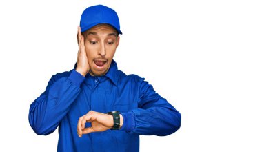 Kel, sakallı, inşaatçı tulumu giyen, saati izleyen, geç kalmaktan korkan bir adam. 