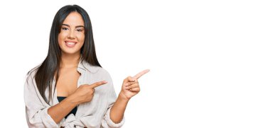 Gündelik giysiler giyen, gülümseyen ve kameraya bakan genç, güzel İspanyol kız iki eliyle ve parmaklarıyla kenarı işaret ediyor.. 