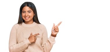 Kışın yün kazak giyen, gülümseyen ve kameraya bakan genç Latin kız iki elini ve parmağını kenarı işaret ediyor.. 