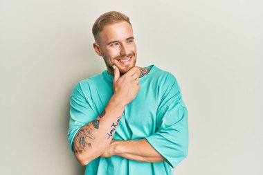 Elleri çenesinde gündelik kıyafetler giyen genç beyaz bir adam soru soruyor, düşünceli bir yüz ifadesi var. Düşünceli bir yüzle gülümsüyor. şüphe kavramı. 