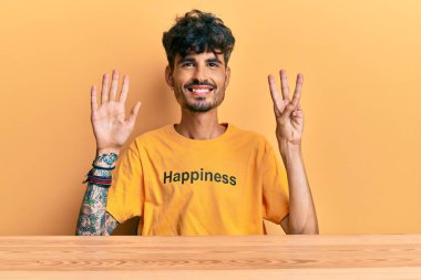 Üzerinde mutluluk mesajı olan İspanyol tişörtü giyen genç adam masanın üzerinde oturuyor ve sekiz numaralı parmaklarıyla işaret ediyor kendinden emin ve mutlu gülümserken.. 