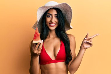 Güzel İspanyol kadın bikini giyiyor, karpuz yiyor gülümsüyor, mutlu bir şekilde elini ve parmağını yana doğru gösteriyor. 