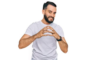 Sakallı, sıradan beyaz tişörtler giyen genç adam ellerini kavuşturup gülümseyerek gevşemiş ve neşeli bir şekilde dua ediyor. Başarı ve iyimserlik 