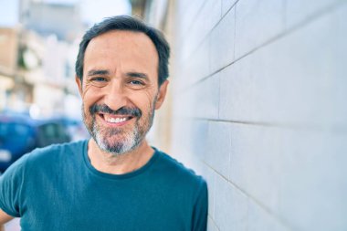 Orta yaşlı, sakallı, mutlu, açık havada duvara yaslanmış bir adam.