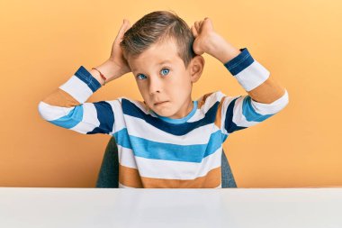 Kafkasyalı sevimli bir çocuk, gündelik giysiler giyip masaya oturmuş, tavşan kulağı hareketi yapıyor, avuç içleri alaycı ve şüpheci görünüyor. Paskalya tavşanı konsepti. 