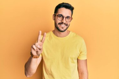 Gündelik kıyafetler giyen ve gözlüklü genç İspanyol adam iki numarayı gösterip işaret ederken kendinden emin ve mutlu bir şekilde gülümsüyor.. 