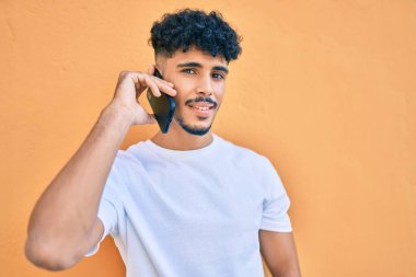 Genç Arap adam gülümsüyor. Akıllı telefonuyla konuşuyor. Duvara yaslanıyor..