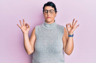 Artı, sıradan kıyafetler ve gözlük takan esmer kadın rahatla ve gözün kapalı gülümse parmaklarınla meditasyon hareketi yap. yoga konsepti. 