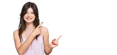 Gündelik kıyafetler giyen, gülümseyen ve kameraya bakan genç, güzel beyaz bir kız. İki eliyle ve parmaklarıyla kenarı işaret ediyor.. 