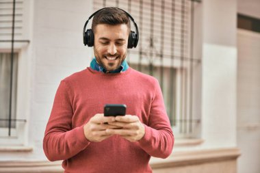 Genç İspanyol adam şehirde akıllı telefon ve kulaklık kullanarak mutlu gülümsüyor..