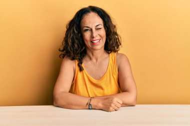 Güzel orta yaşlı kadın gündelik giysiler içinde masaya oturmuş kameraya seksi bir ifadeyle bakıyor, neşeli ve mutlu bir yüzle.. 