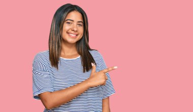 Yüzünde mutlu ve doğal bir ifade olan, elleri ve parmaklarıyla işaret eden, güler yüzlü, gündelik kıyafetler giyen genç Latin kız. 