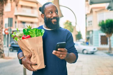 Sakallı Afrikalı Amerikalı adam elinde süpermarketten alınmış kağıt torbalarla akıllı telefon kullanıyor.