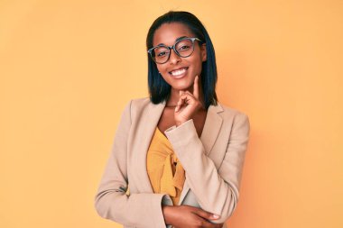Genç Afro-Amerikan kadın iş kıyafetleri giyiyor kameraya özgüvenli bakıyor kollarını kavuşturup gülümseyerek ve çenesini kaldırarak. Olumlu düşünmek. 