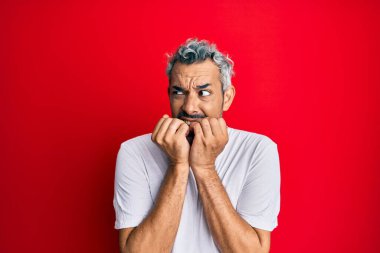 Orta yaşlı, beyaz tişört giyen, gergin ve gergin görünen, elleri ağızdan tırnak yiyen gri saçlı bir adam. anksiyete. 