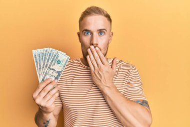Dolarları tutan genç beyaz bir adam ağızlarını eliyle kapatıyor, şaşırmış ve hata yapmaktan korkuyor. şaşırmış ifade 