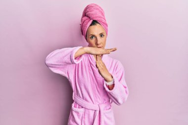 Duş havlusu şapkası ve bornoz takan genç sarışın kadın. El kol hareketi yapıyor, hayal kırıklığına uğramış ve ciddi bir yüz ifadesi var. 