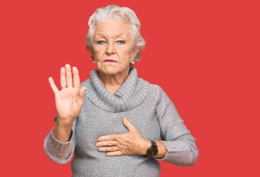 Kış süveteri giyen, elleri göğsünde ve avuçlarında küfreden, sadakat yemini eden, kıdemli gri saçlı kadın. 