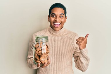 Genç, yakışıklı, İspanyol asıllı, elinde çikolata parçacıklı kurabiye kavanozu olan, başparmağıyla yan tarafa bakan, ağzı açık gülümseyen bir adam. 