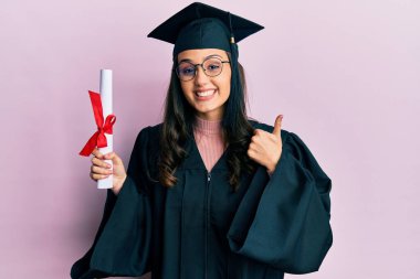 Mezuniyet üniforması giyen genç İspanyol bir kadın. Elinde mutlu ve olumlu bir gülümseme, başparmağında mükemmel ve onay işareti var. 