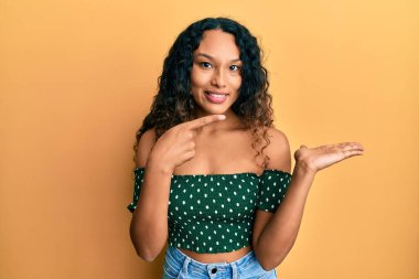 Gündelik kıyafetler giyen genç Latin kadın kameraya hayret ediyor ve gülümsüyor.. 