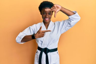 Genç Afrikalı Amerikalı kız karate kimonosu ve siyah kuşak takmış gülümsüyor ve elleri ve parmakları gülen yüzlü bir çerçeve yapıyor. yaratıcılık ve fotoğrafçılık kavramı. 