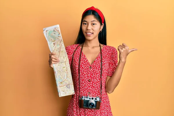 Young chinese woman using city map and vintage camera pointing thumb up to the side smiling happy with open mouth