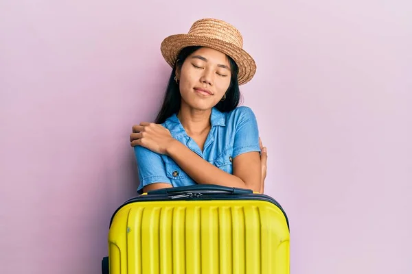 Young chinese woman wearing summer hat and holding cabin bag hugging oneself happy and positive, smiling confident. self love and self care