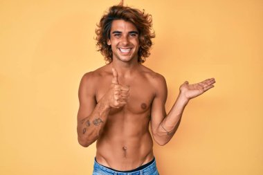 İspanyol asıllı genç adam avucunu gösteriyor ve baş parmaklarıyla iyi bir hareket yapıyor, mutlu ve neşeli gülümsüyor. 