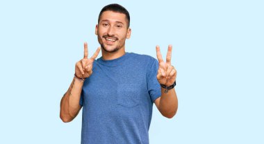 Dövmeleri olan, sıradan kıyafetler giyen, gülümseyen, parmaklarını zafer işareti yaparken gösteren yakışıklı bir adam. İki numara.. 