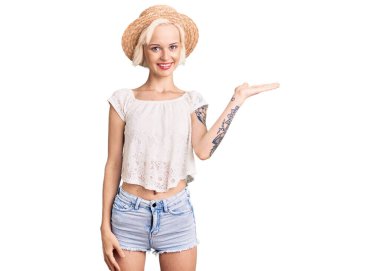 Yazlık şapkalı, neşeli gülümseyen, avuç içiyle kameraya bakan sarışın genç bir kadın.. 