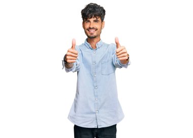 Latin asıllı genç adam gündelik giysiler giyip olumlu jestler yapıyor. Elleriyle gülümsüyor ve başarı için seviniyor. kazanan jest. 