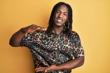 Saçları örgülü, leopar desenli hayvan desenli tişörtlü, elleri büyük ve büyük bir işaret gösteren Afro-Amerikan bir adam. Kameraya bakıp gülümsüyor. ölçüm kavramı. 