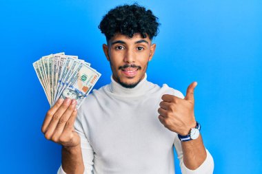 Genç Arap yakışıklı adam elinde dolarlarla mutlu ve pozitif gülümsüyor, başparmağıyla mükemmel ve onay işareti yapıyor. 
