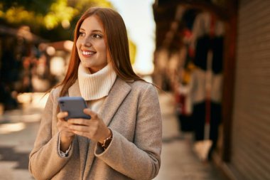 Genç kızıl saçlı kız şehirde akıllı telefon kullanarak mutlu bir şekilde gülümsüyor..