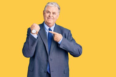 Kıdemli gri saçlı adam iş ceketi giyiyor. Aceleyle saati, sabırsızlığı ve rahat bir ifadeyle kameraya bakıyor. 