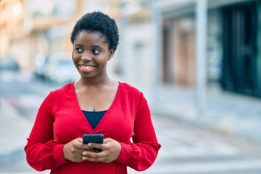 Genç Afrikalı Amerikalı kadın şehirde akıllı telefon kullanarak mutlu bir şekilde gülümsüyor..