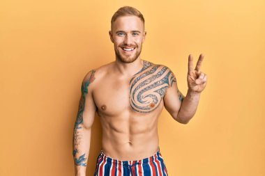 Mayo giyen genç, beyaz bir adam iki numarayı gösterip kendini beğenmiş ve mutlu bir şekilde gülümsüyor.. 