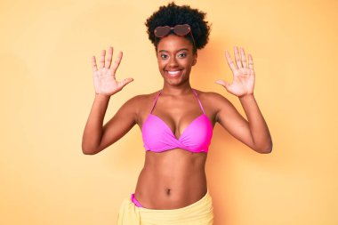Genç Afro-Amerikan kadın bikini giyiyor ve parmaklarıyla on numarayı gösterirken kendinden emin ve mutlu gülümsüyor.. 