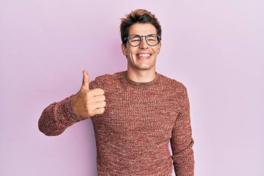 Yakışıklı beyaz adam, günlük süveter giyiyor ve gözlük takıyor. Elle mutlu bir jest yapıyor. Kameraya bakıp başarıyı gösteren ifade. 