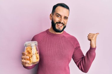 Elinde makarna kavanozu olan sakallı genç bir adam başparmağını yukarı kaldırıyor ve ağzı açık gülümsüyor. 