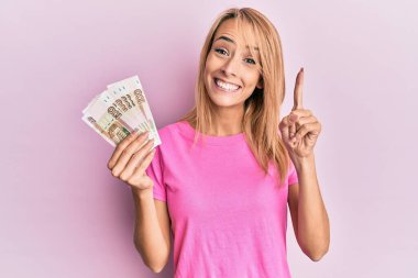Güzel sarışın kadın elinde 100 Rus rublesi banknotuyla bir fikir veya soruyla gülümsüyor ve mutlu bir yüzle işaret ediyor. 
