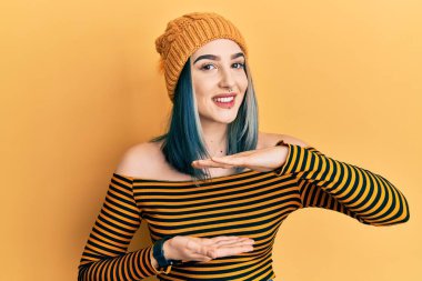 Yün şapka takan genç bir kız. Elleriyle işaret ediyor. Büyük ve büyük beden işareti, ölçü işareti. Kameraya bakıp gülümsüyor. ölçüm kavramı. 