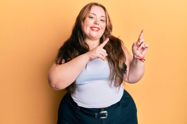 Artı, günlük kıyafetler giyen beyaz bir kadın. Gülümsüyor ve kameraya bakıyor. İki eliyle ve parmaklarıyla onu işaret ediyor.. 